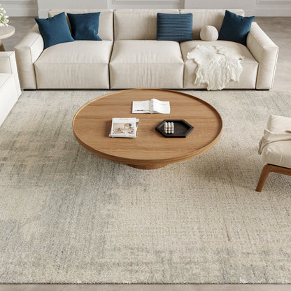 Round wooden coffee table on a pale rug, beige sofa with blue cushions and a white throw; magazines and a black tray on top.
