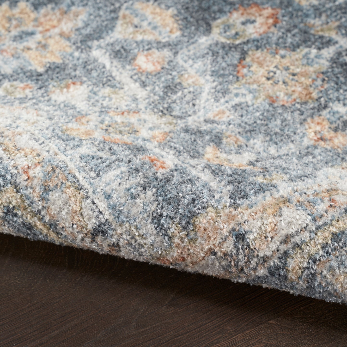 Blue-gray floral patterned plush area rug with beige and orange accents on dark wood floor.
