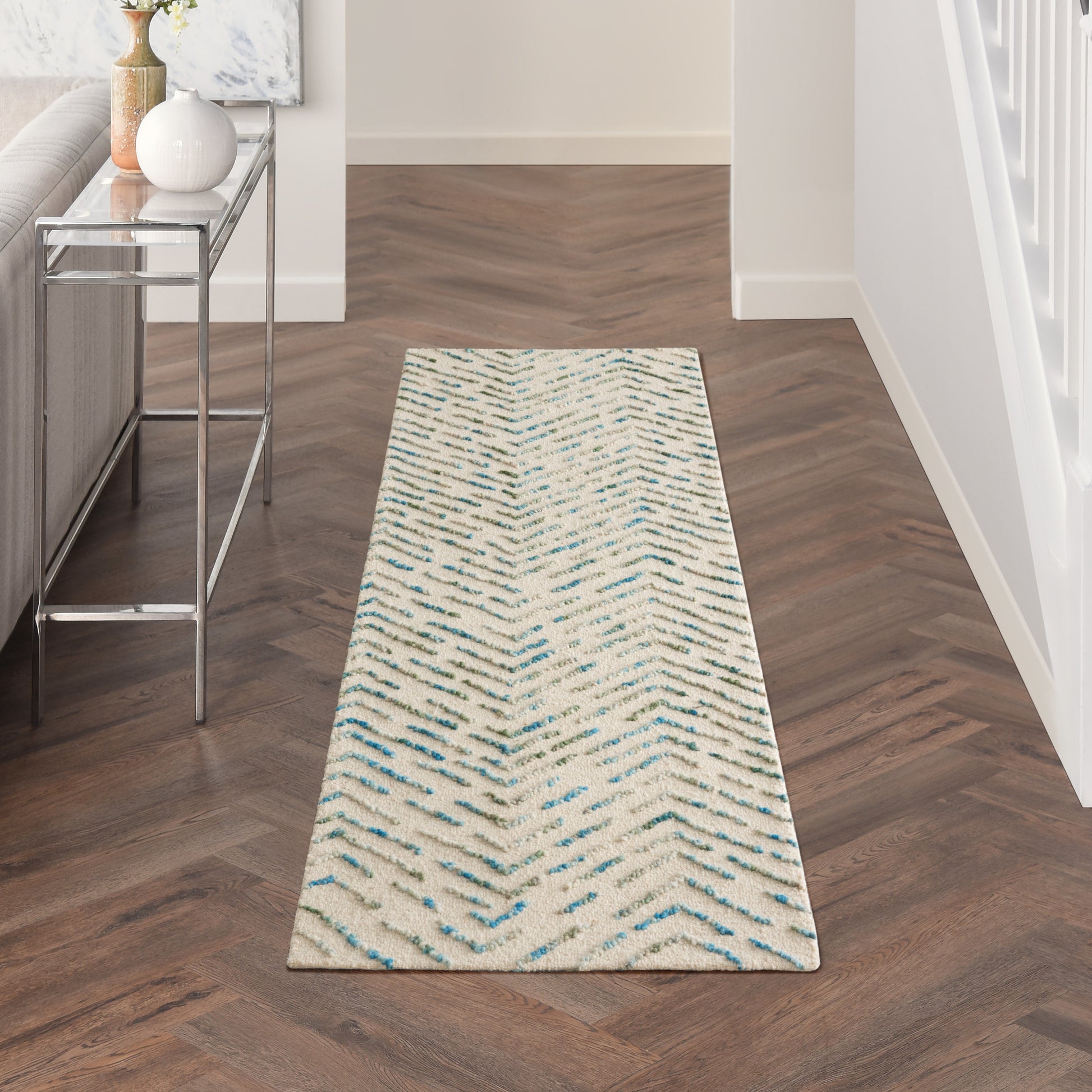 Long beige hallway runner rug with blue chevron pattern on dark hardwood floor beside a glass-top metal console.