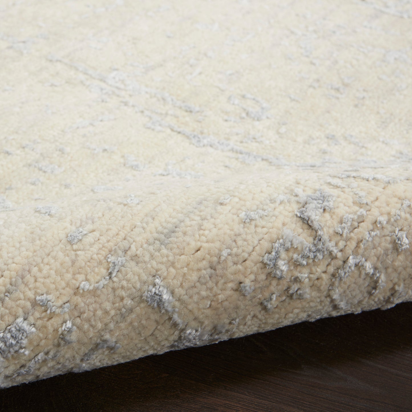 Beige plush area rug with a soft pile and subtle light gray abstract pattern, edge curled on dark wooden floor.