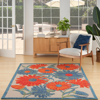 Rectangular woven outdoor rug with bright orange and blue floral pattern on beige, teal border; shown in a light-wood outdoor setting, 6' x 9'.