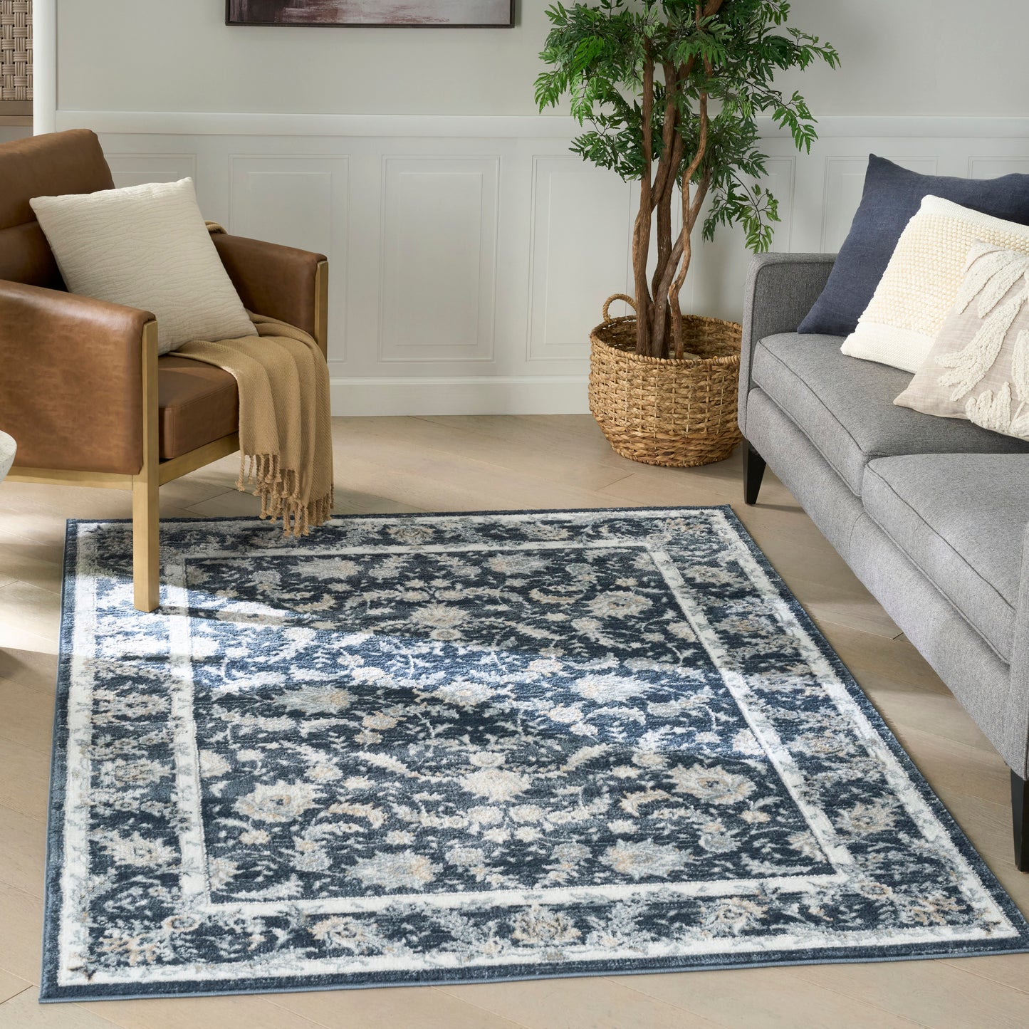 Blue and cream ornate area rug in a living room with gray sofa, brown leather chair, plant in wicker basket, wood floor.