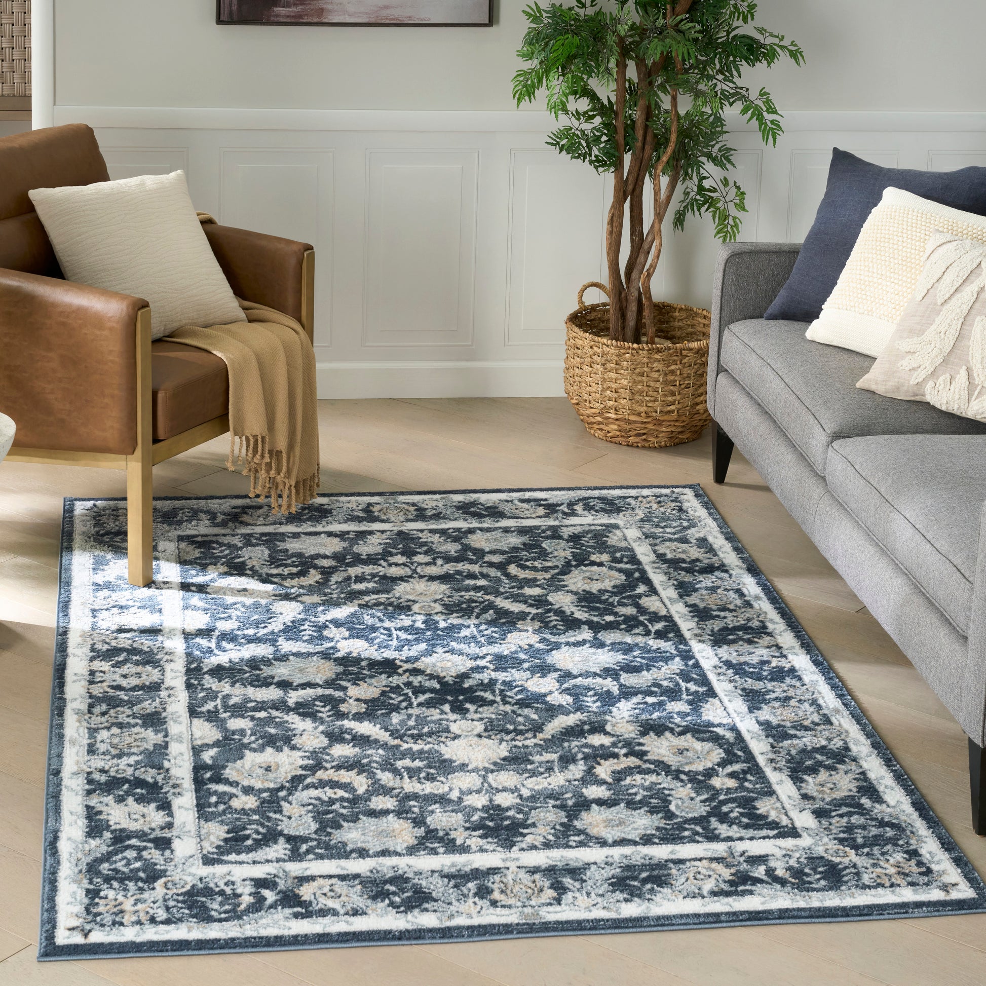 Blue and cream ornate area rug in a living room with gray sofa, brown leather chair, plant in wicker basket, wood floor.