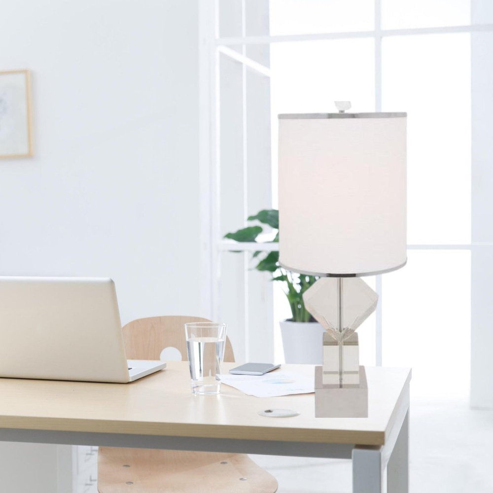 Light wood desk with laptop, glass of water, and a modern geometric table lamp; bright window and a plant in the background.