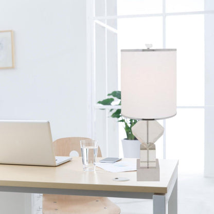 Light wood desk with laptop, glass of water, and a modern geometric table lamp; bright window and a plant in the background.
