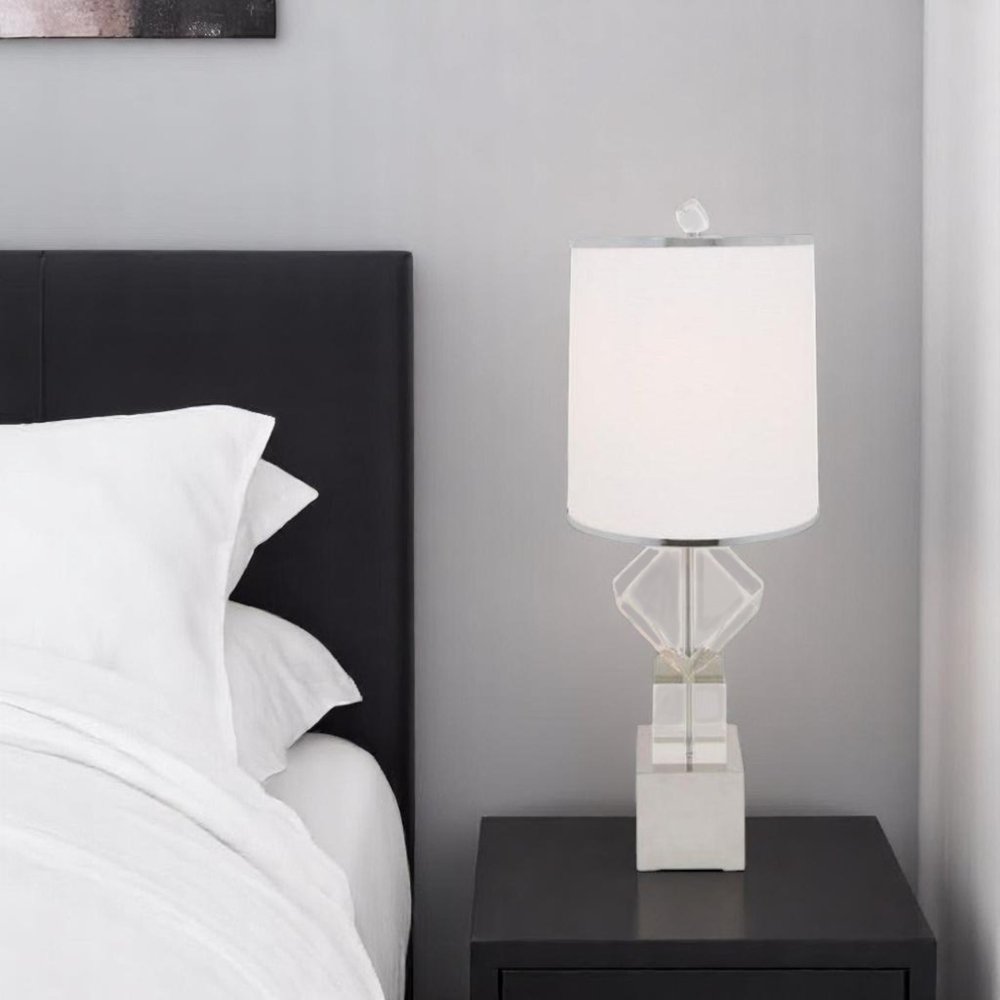 Modern geometric glass table lamp with white shade on a black nightstand beside a white bed against a gray wall.