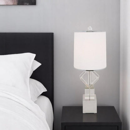 Modern geometric glass table lamp with white shade on a black nightstand beside a white bed against a gray wall.