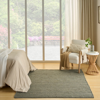 Neutral bedroom with beige bedding, gray rug, white lounge chair, round wooden side table with a potted plant, by large glass doors.