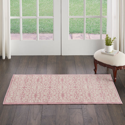 Pink damask rug with pale border on dark wood floor, beside a cream stool with plant near glass doors.