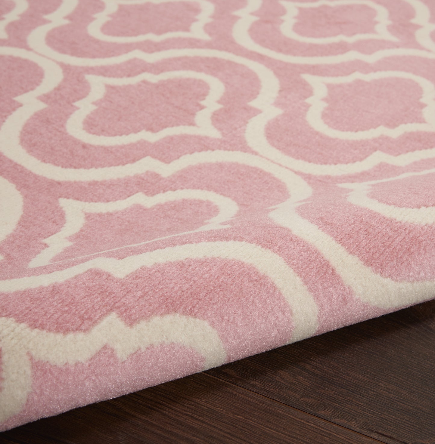 Soft pink area rug with a white abstract trellis pattern on dark wood floor.