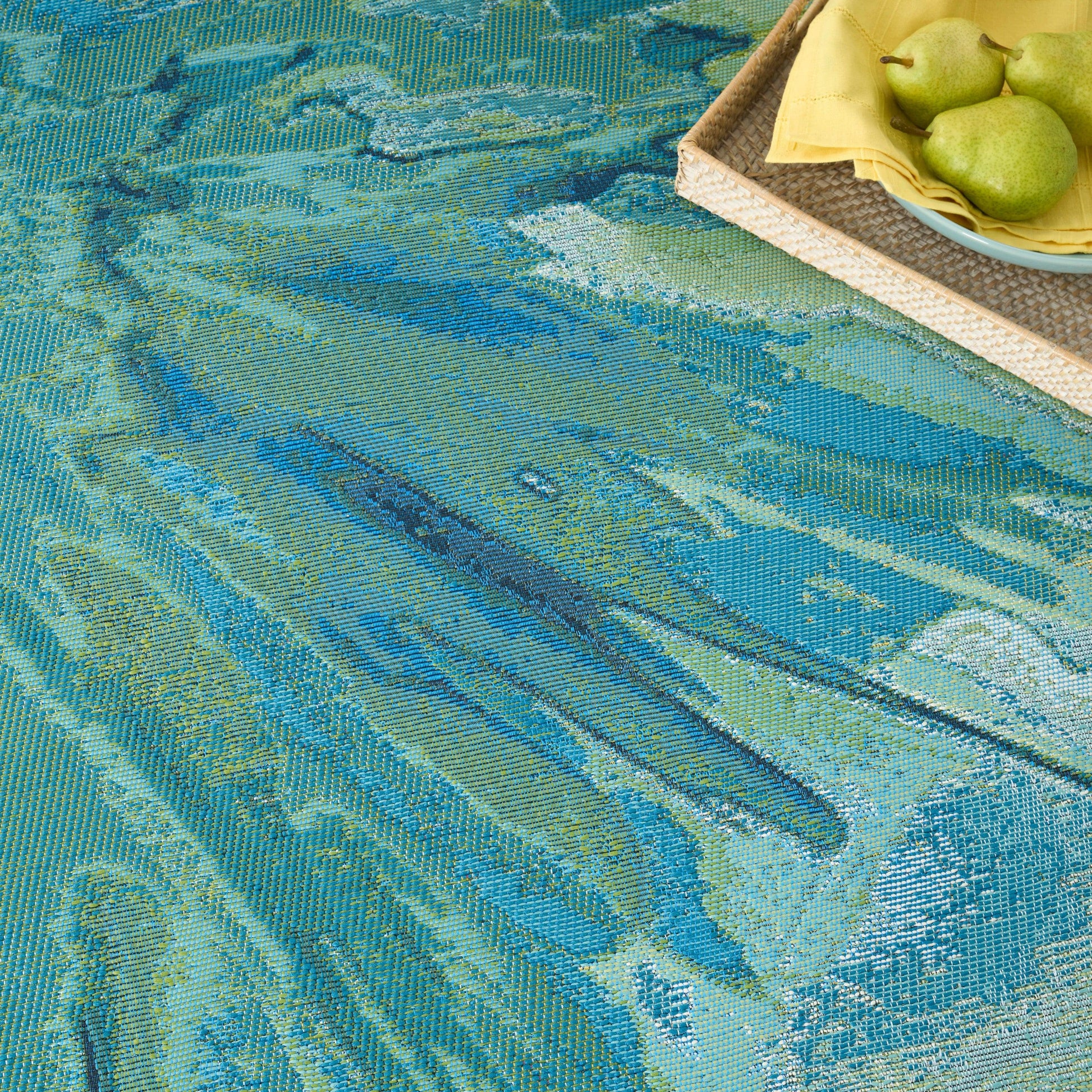 Rectangular teal, blue and green abstract woven rug with a wicker tray, a yellow napkin and green pears.