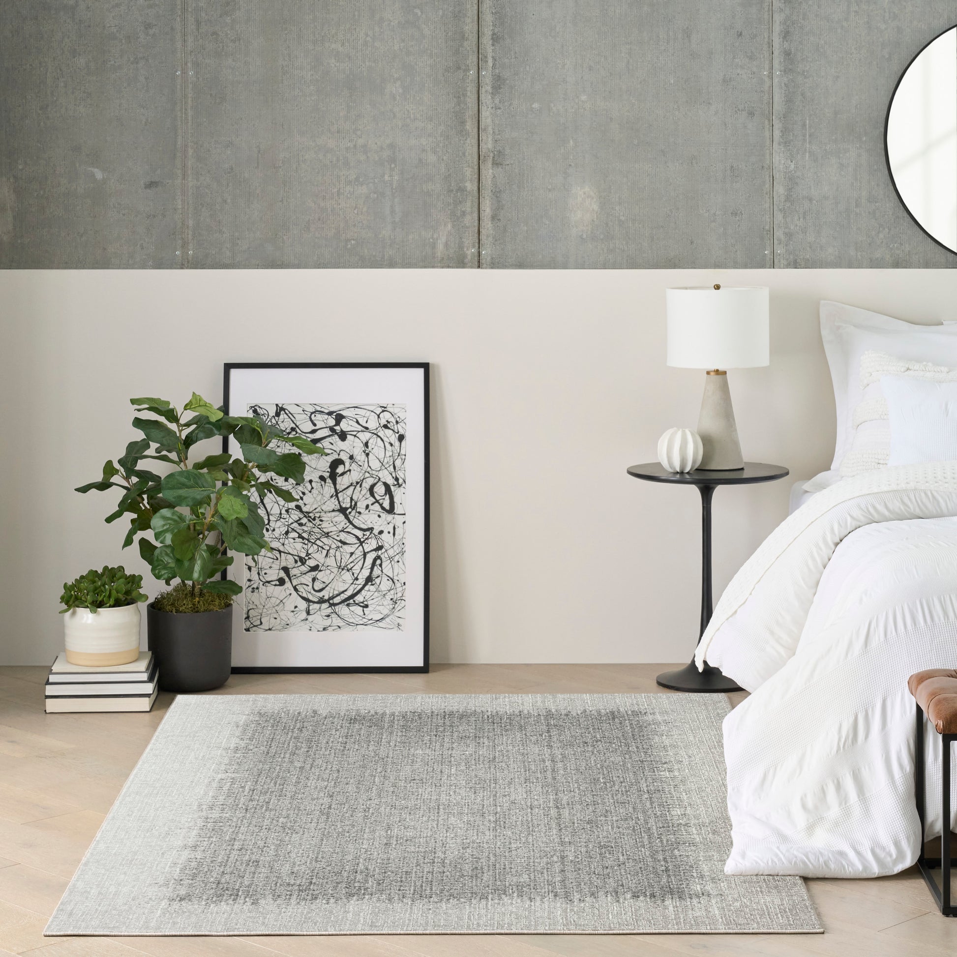 Minimalist bedroom: white bedding, black-framed abstract art, potted plant, books, round side table, lamp, light gray rug.