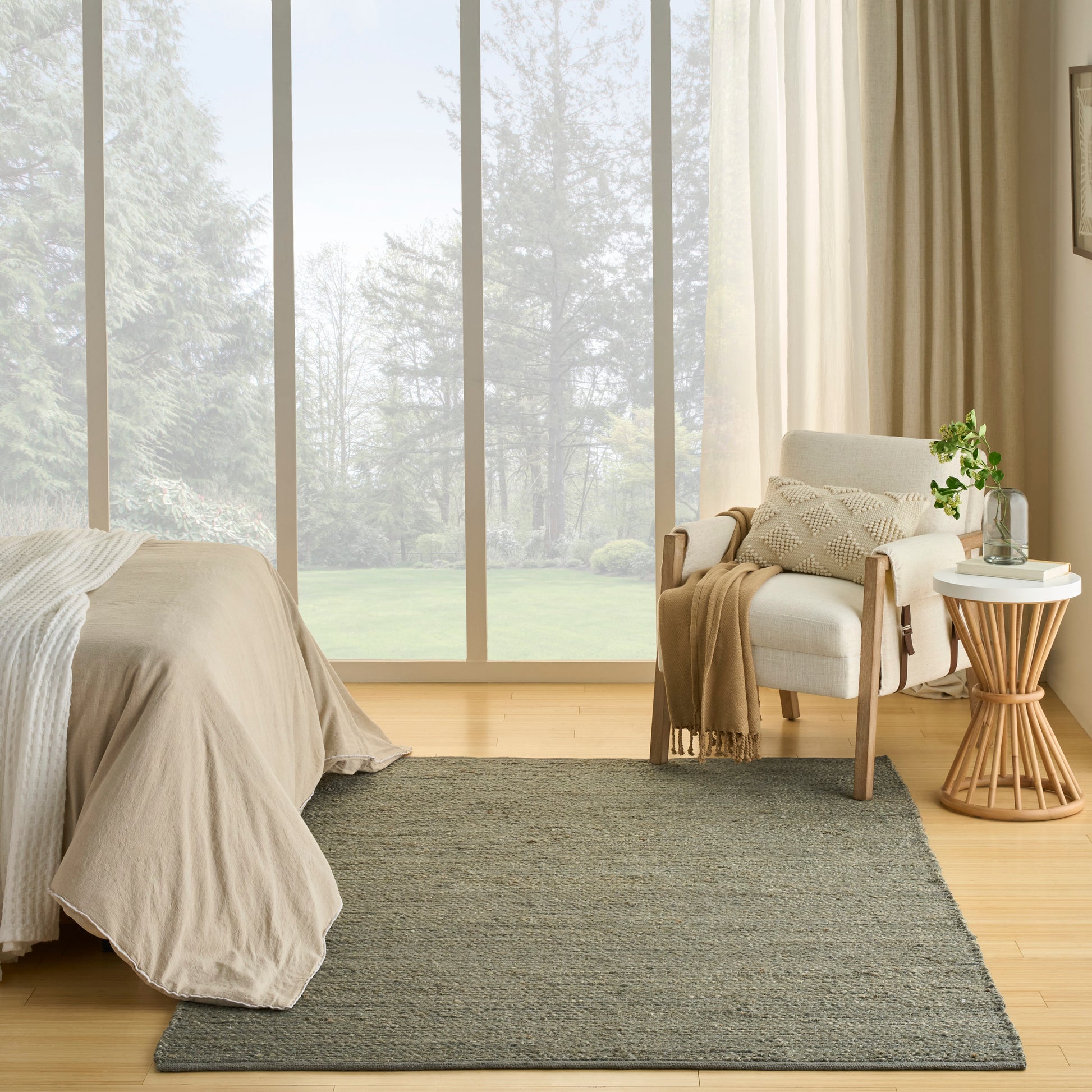 Neutral bedroom with beige bedspread, gray rug, armchair, round wooden side table, potted plant, and large windows.