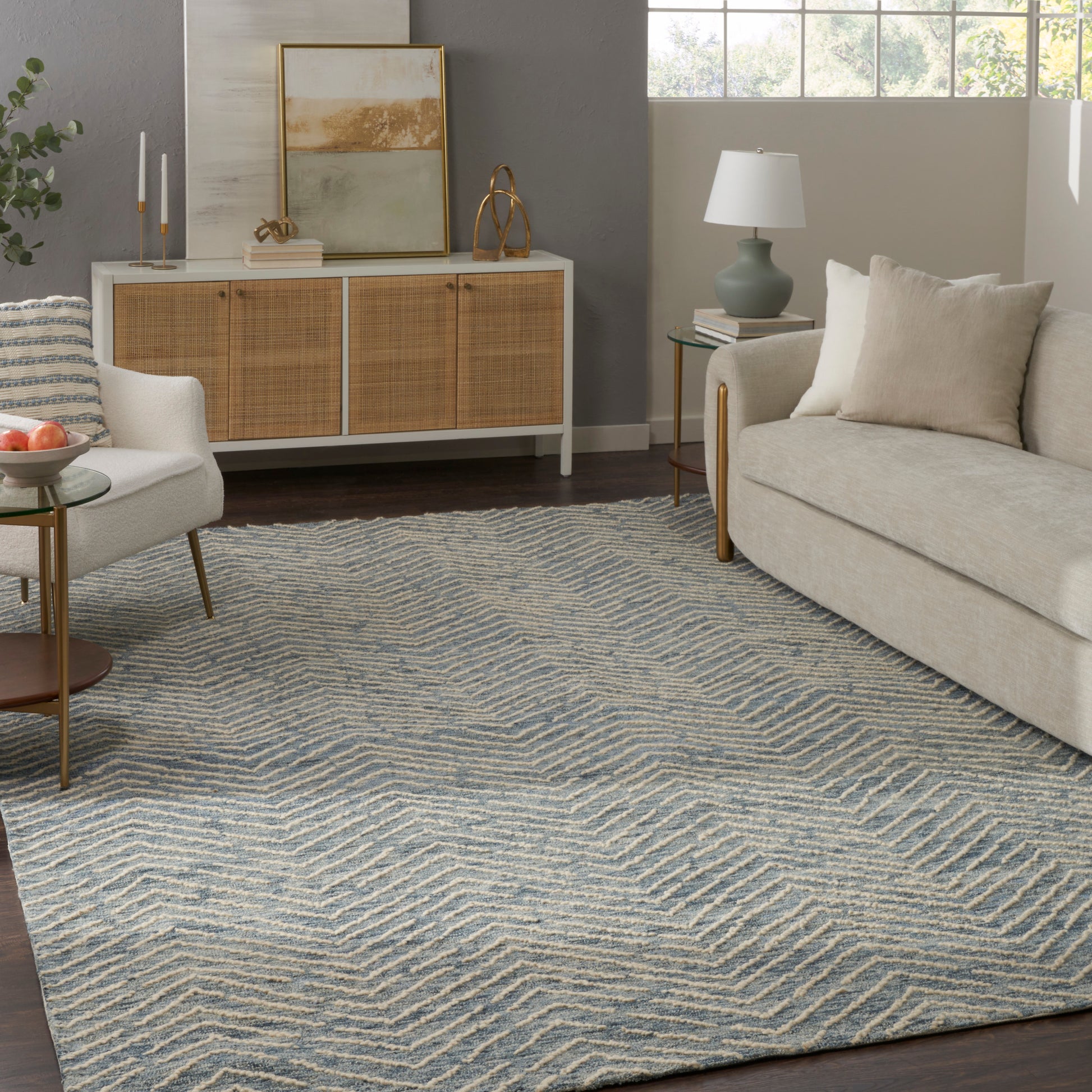 Textured blue and beige chevron area rug in a modern coastal living room with a beige sofa, glass coffee table, and rattan cabinet.