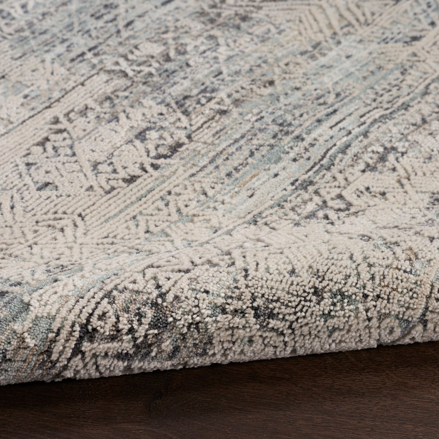 Soft blue-gray patterned area rug with geometric motifs and a plush raised pile; edge visible on dark wood floor.