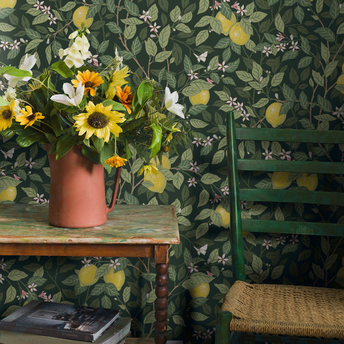 Terracotta vase with sunflowers & white lilies on a weathered wooden table beside a green chair on lemon-patterned wallpaper.