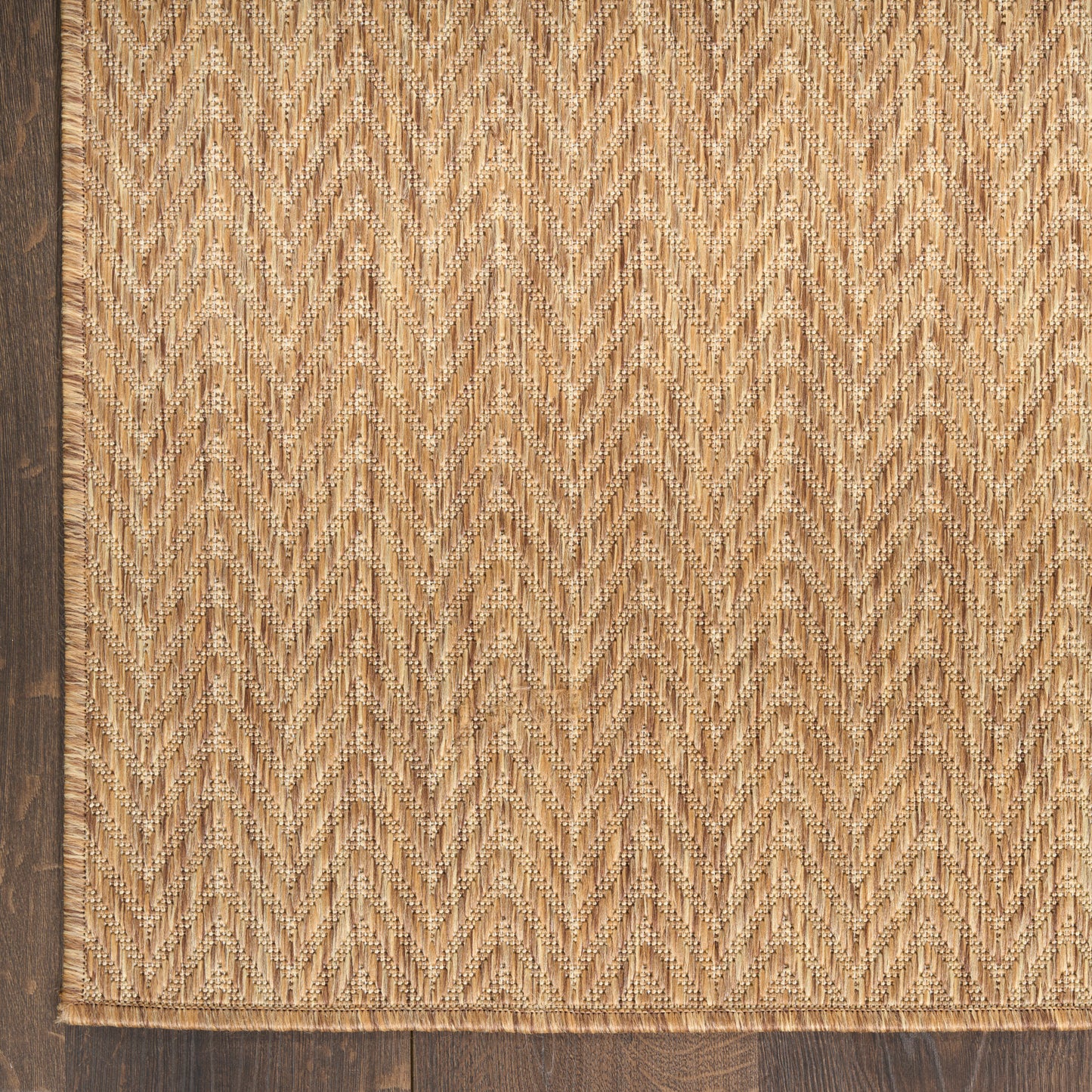 Natural jute rug with chevron/herringbone pattern in beige on dark wood floor, fringed edges.