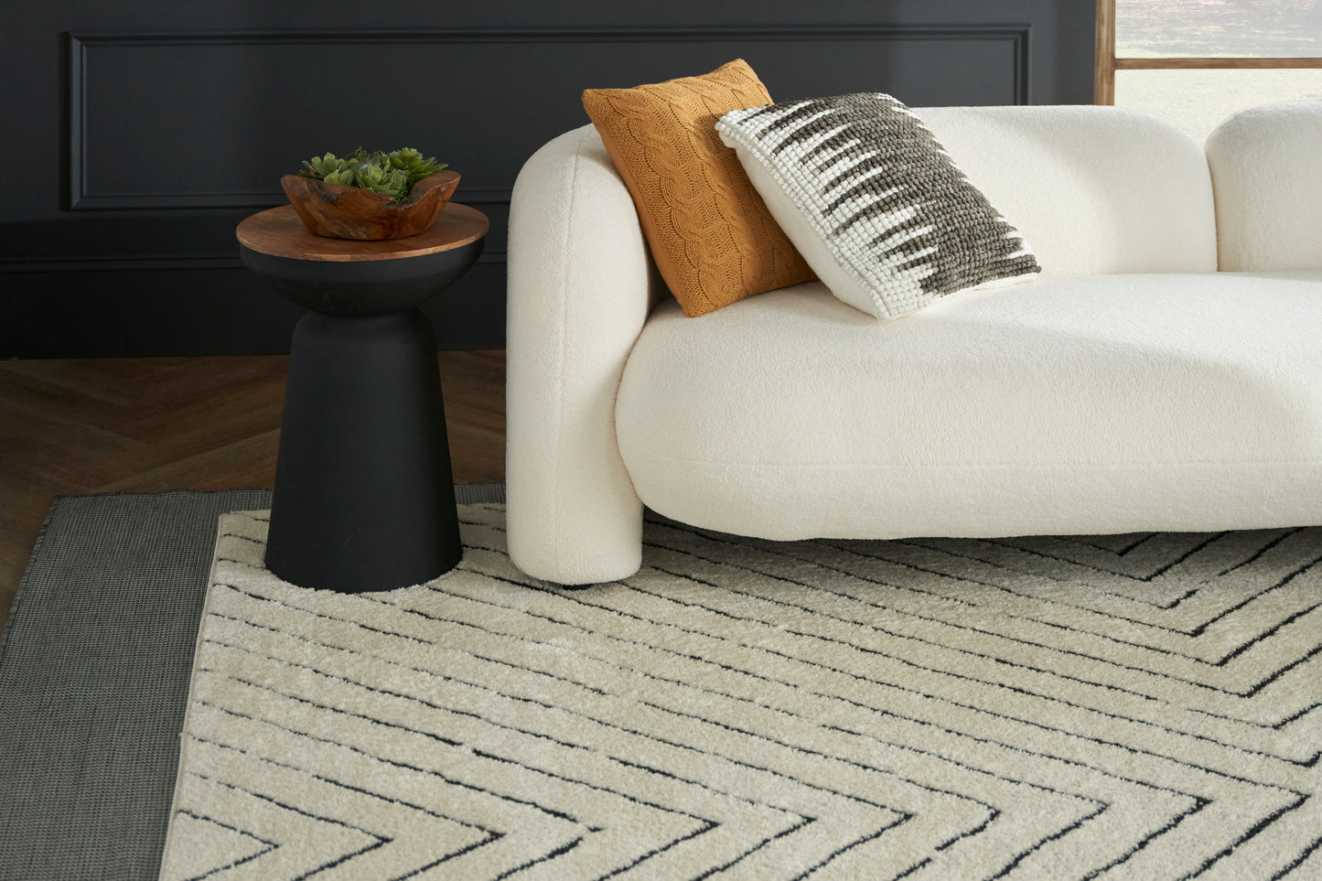 Cream textured area rug with black chevron pattern under a white boucle sofa; mustard throw pillow & black pedestal table.
