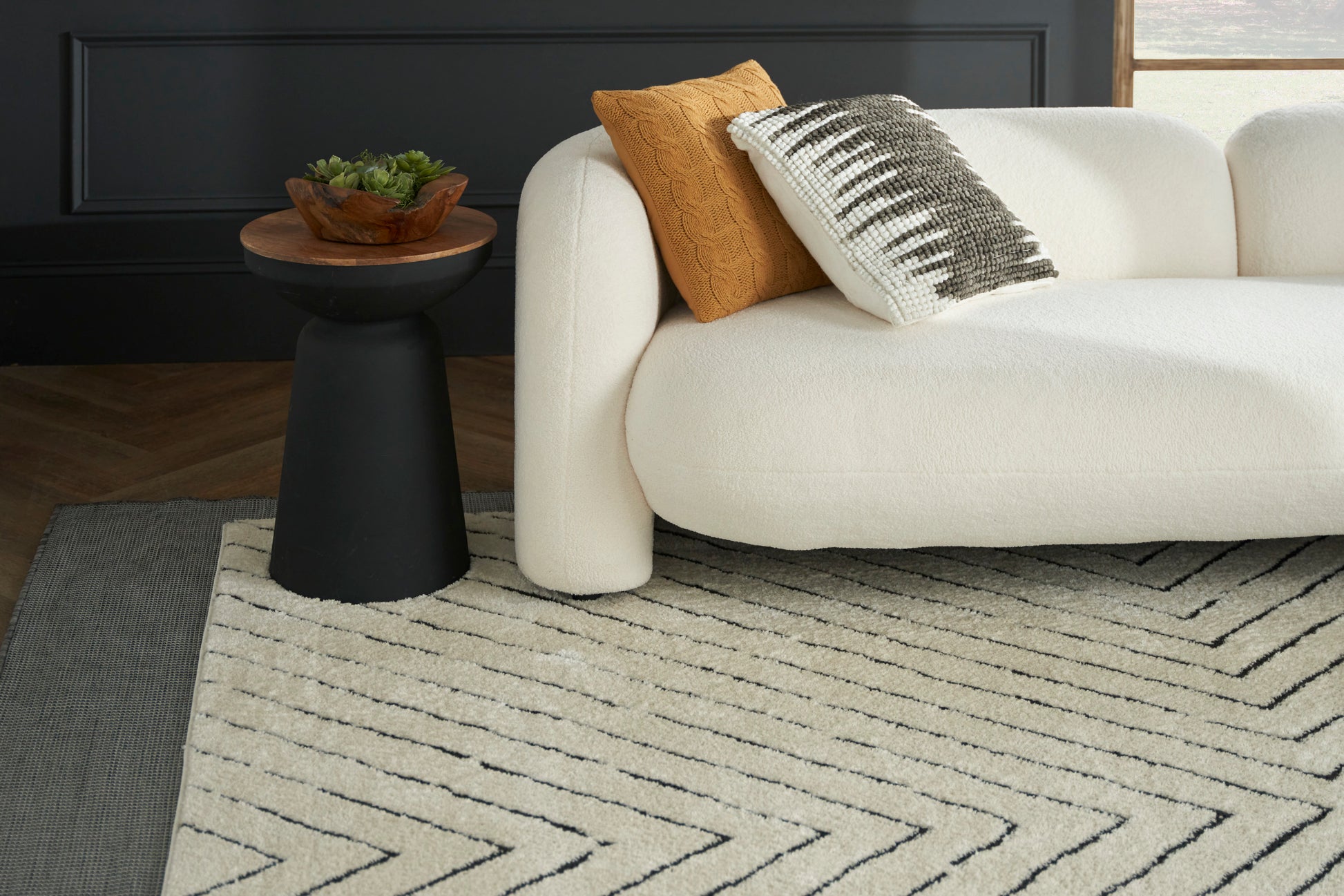 Cream textured area rug with black chevron pattern under a white boucle sofa; mustard throw pillow & black pedestal table.