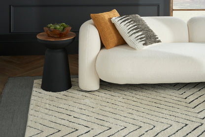 Cream textured area rug with black chevron pattern under a white boucle sofa; mustard throw pillow & black pedestal table.