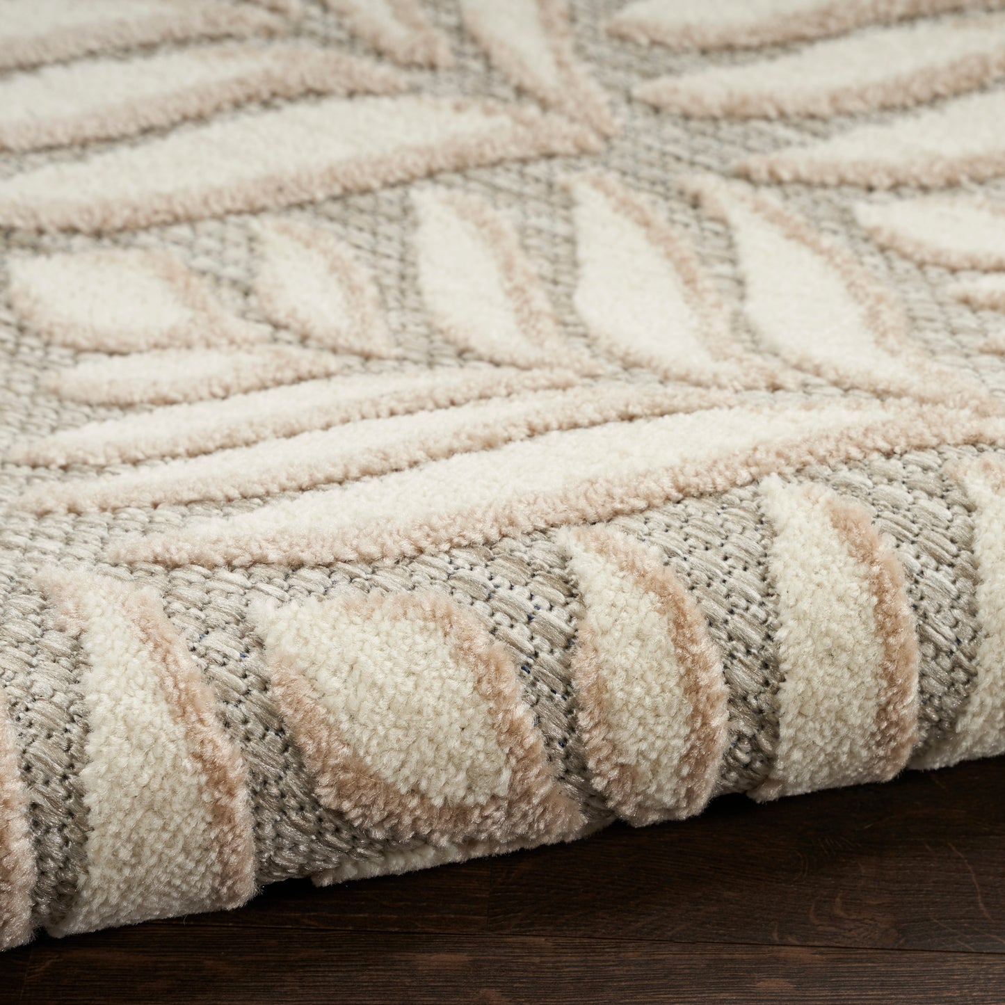 Close-up of a plush beige and gray area rug with a raised leaf pattern and rolled edge on a dark wood floor.