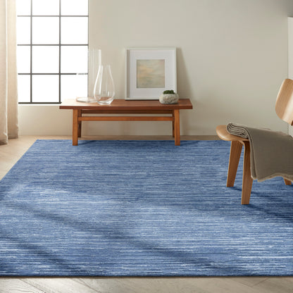 Blue textured area rug in a sunlit modern living room with a low wooden coffee table, glass vases, art, plant, and a chair.