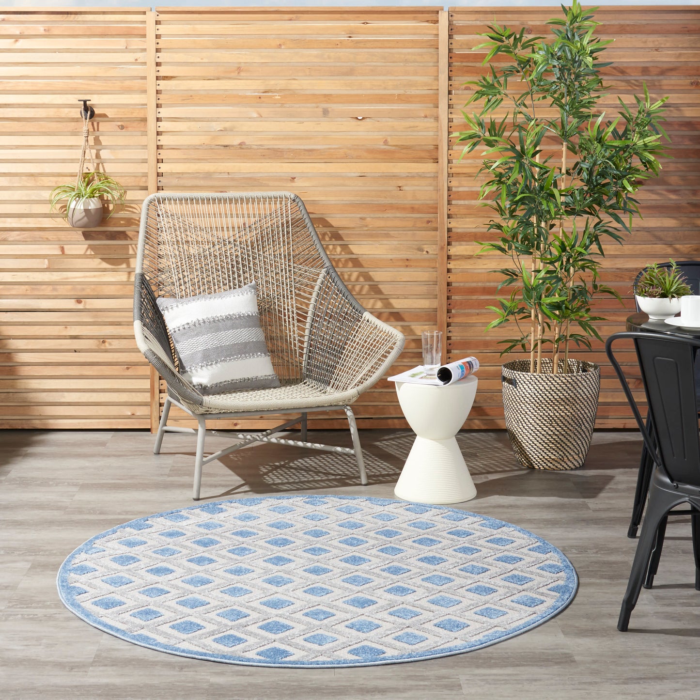 Outdoor patio with beige wicker lounge chair, striped cushion, blue patterned rug, white side table, and potted plants.