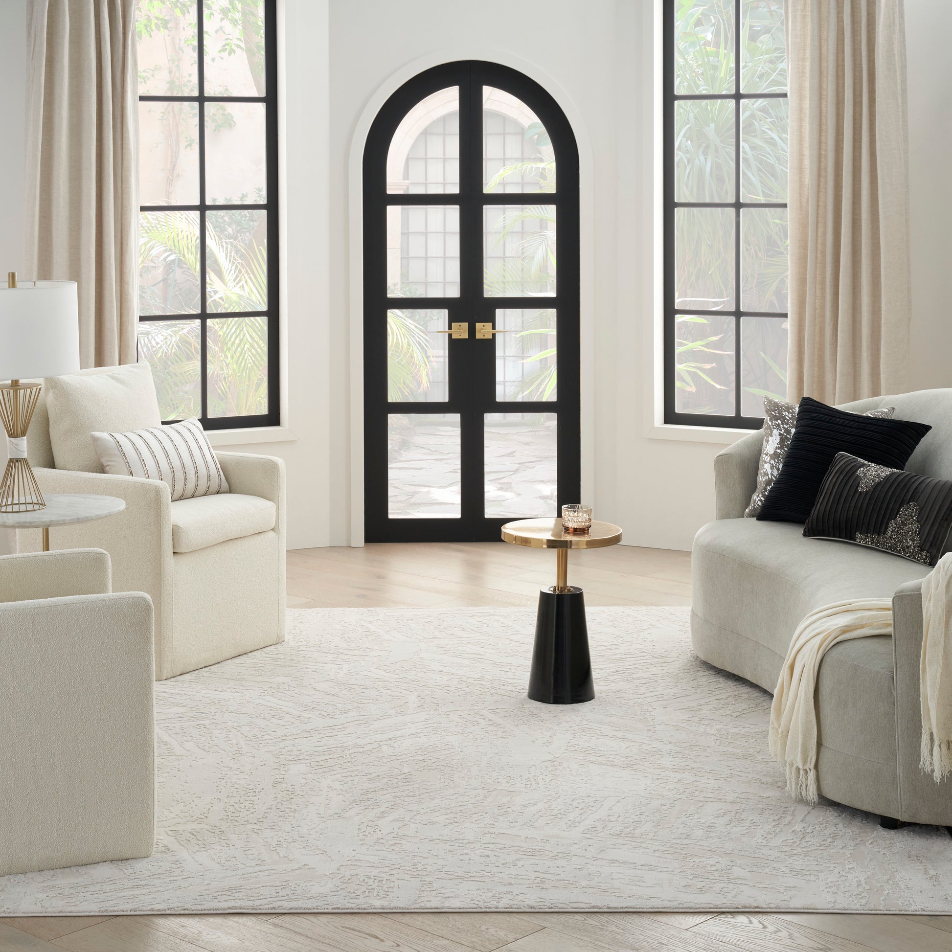 Bright living room with cream sofas, an arched black-framed glass door, large windows, and a gold-and-black round side table.