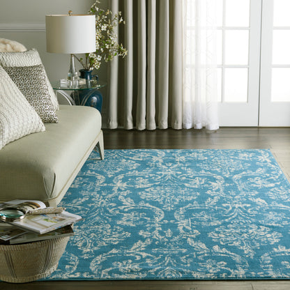 Turquoise blue area rug with white damask pattern on dark wood floor, in a living room with beige sofa and glass side table.
