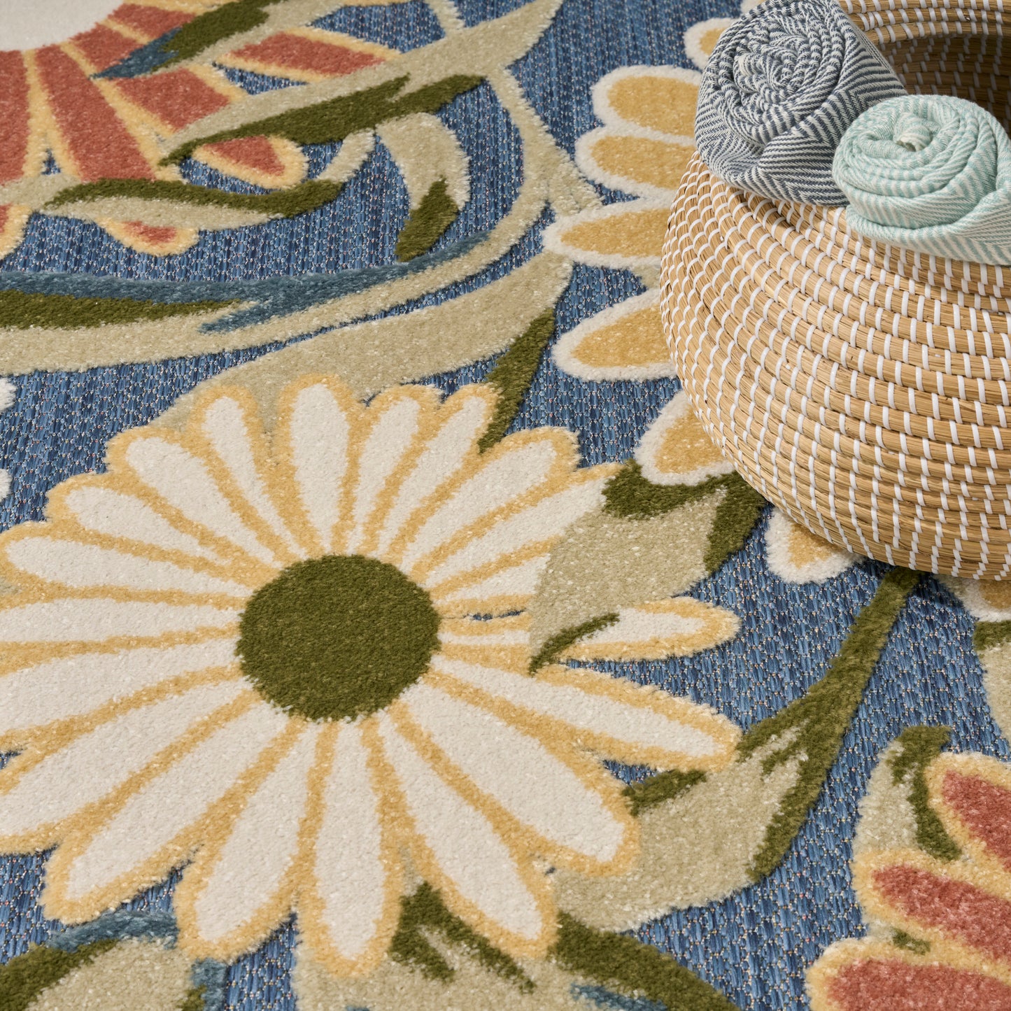 Round woven straw basket with gray and mint fabric rolls on a blue rug with large white and yellow daisies.