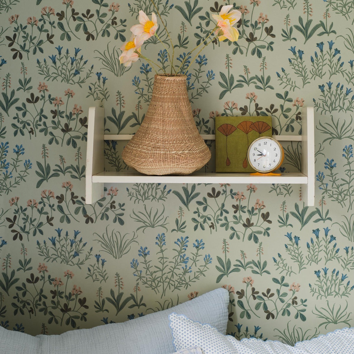 Woven wicker vase with yellow flowers on a white shelf, paired with a vintage clock and green box against wallpaper.