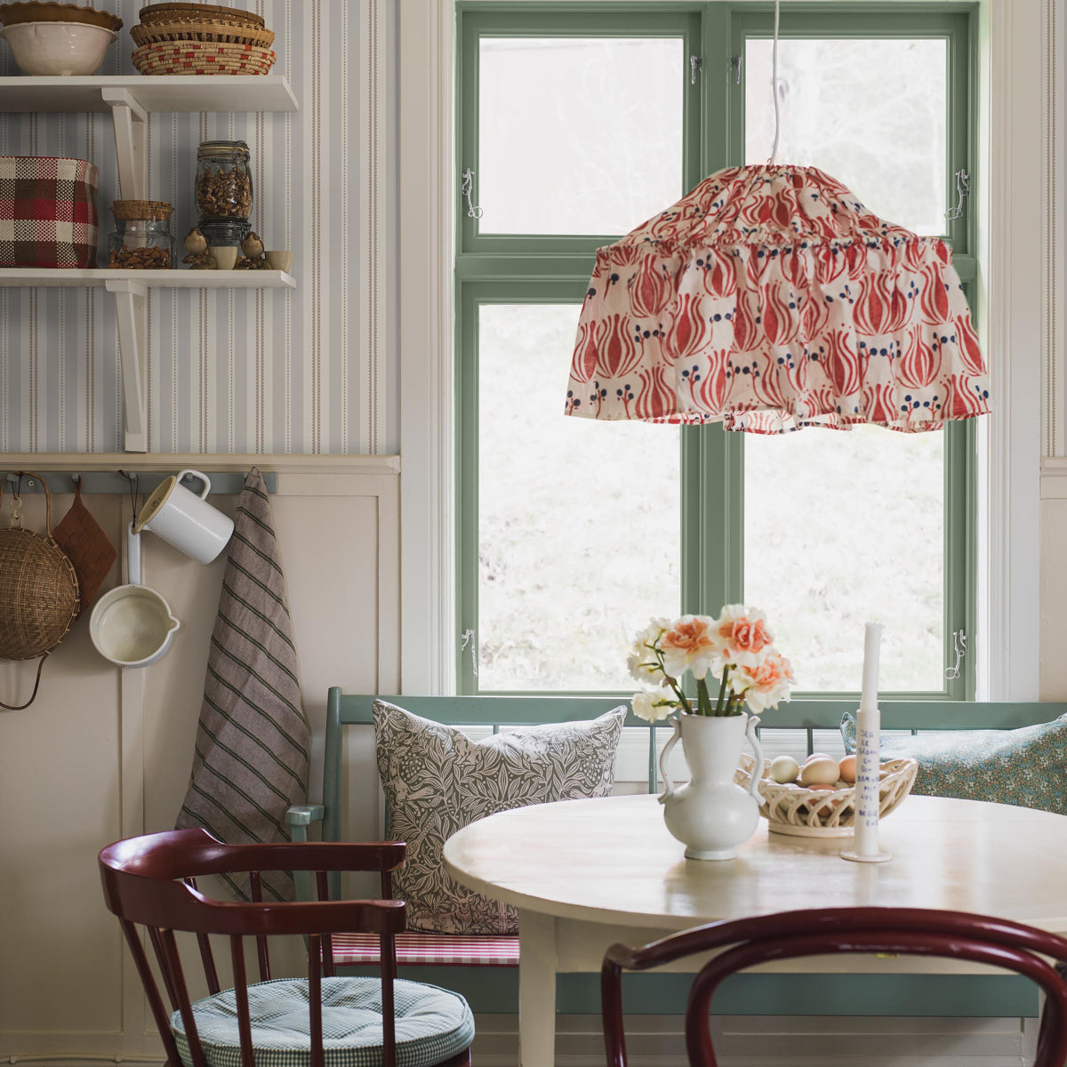 Dining nook with stone taupe wallpaper on walls; round white table, burgundy chairs, green-framed windows, floral red lampshade, and a vase of peaches.
