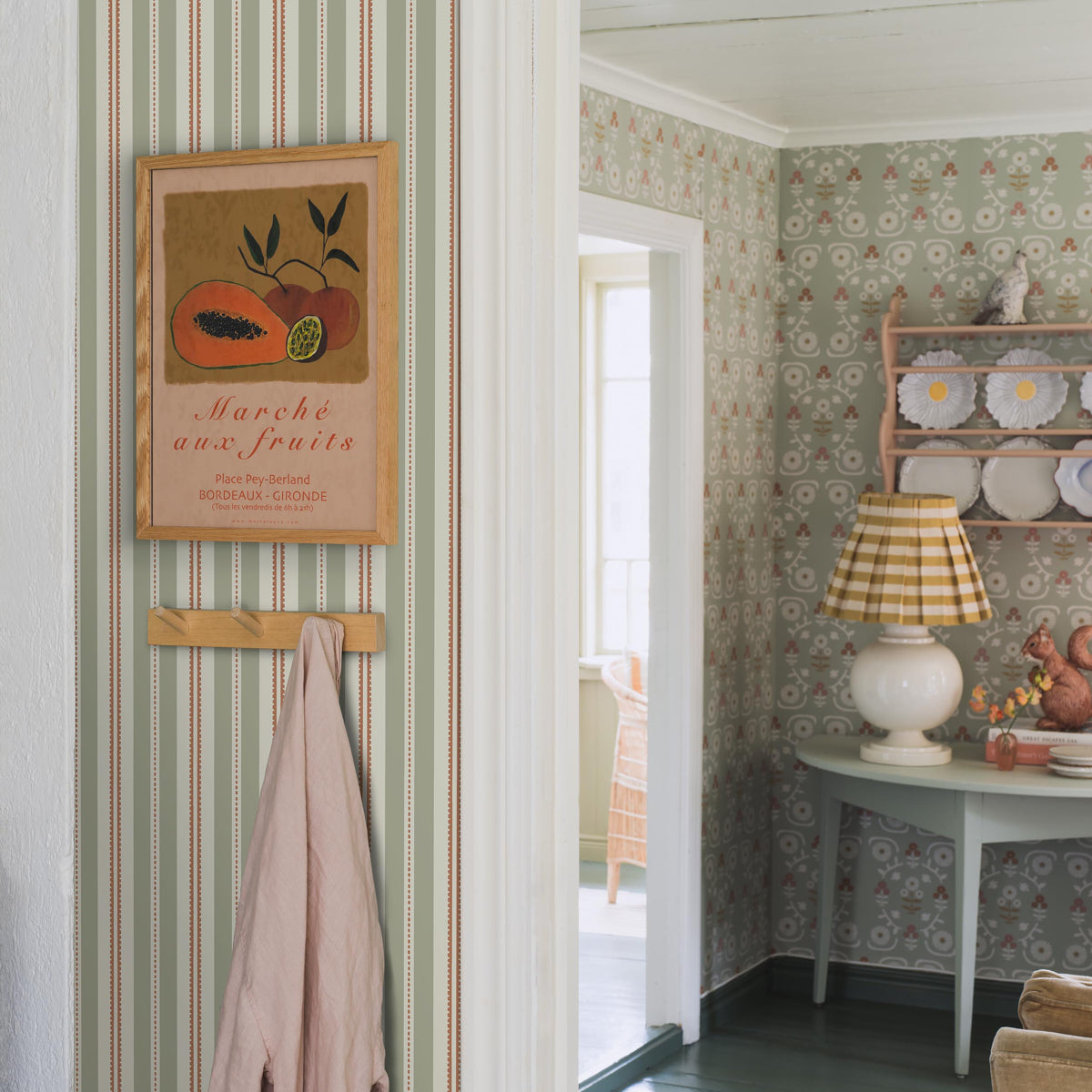 Vintage kitchen corner with green striped wallpaper, framed citrus print, peg rack with towel, and lamp by a dish shelf.