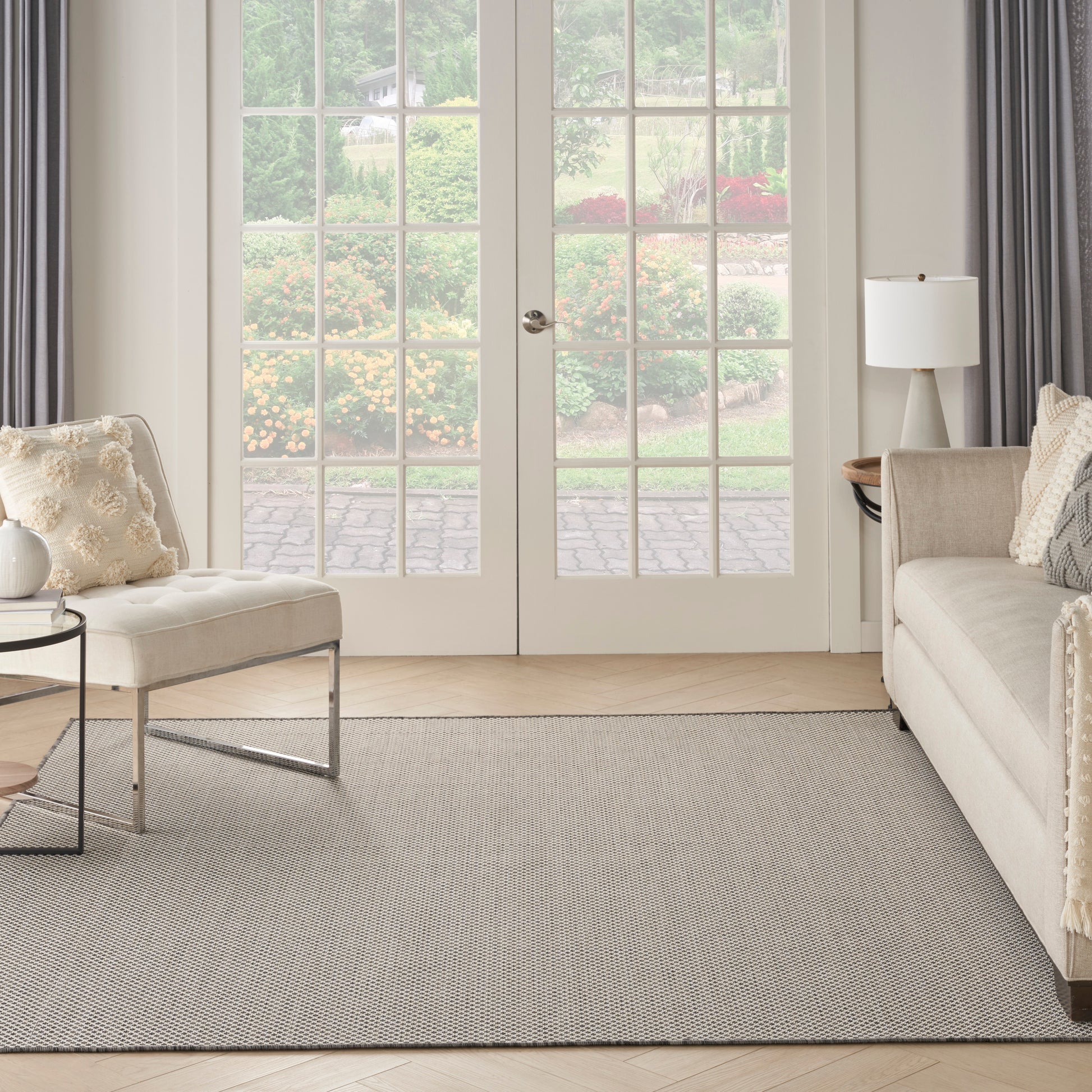Neutral living room with beige sofa and chair, round glass coffee table, gray rug, white lamp, and French doors to garden.