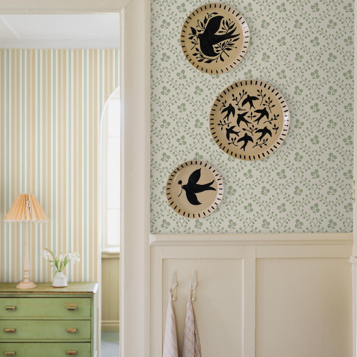 Vintage hallway vignette: green dresser with brass handles, striped and floral wallpaper, and three round wicker plates with black birds.