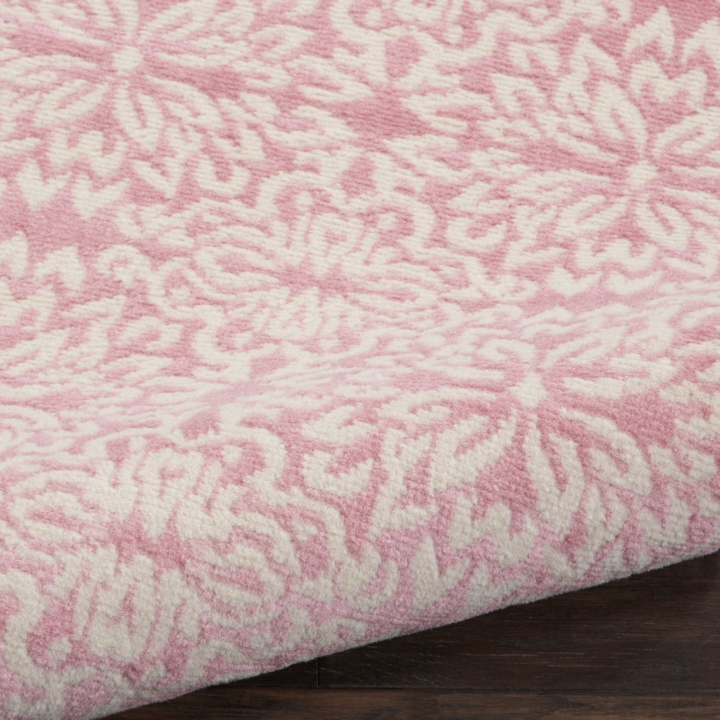 Pink rug with white floral/abstract pattern, soft pile texture, folded edge on dark wood surface.