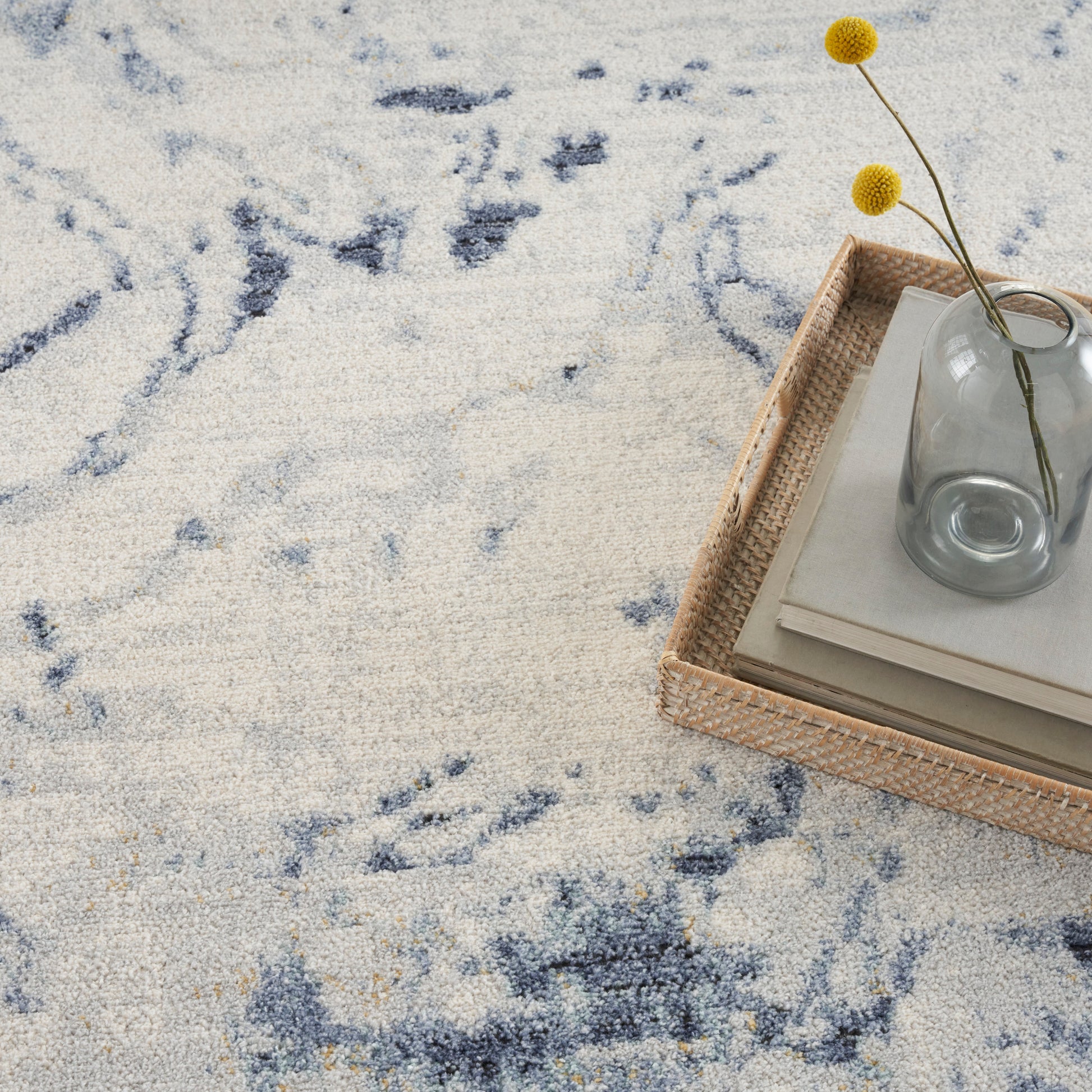 Blue patterned rug 5' x 7' topped with a rectangular woven rattan tray, a glass vase, and yellow billy-ball stems arranged on a stack of books.