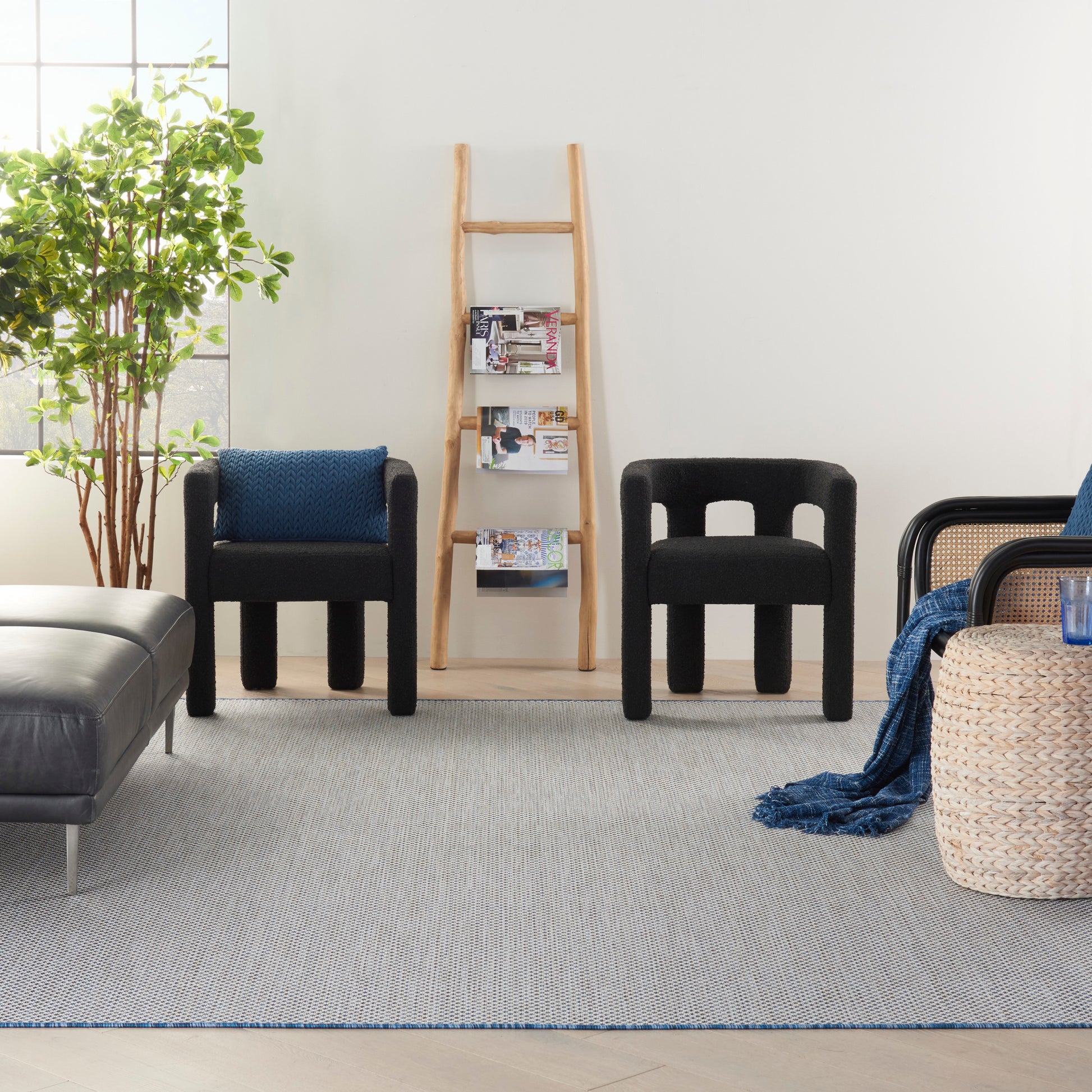 Modern living room with black armless chairs, a wooden ladder shelf, tall plant, grey rug, and a woven basket with blue throw.