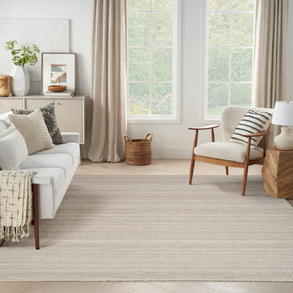 Bright living room with a light beige sofa, cream armchair, woven rug, wooden side table, basket, and large windows.