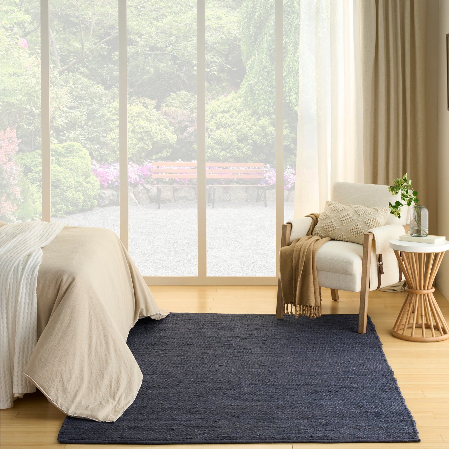 Cozy minimalist bedroom with navy rug, beige bedding, light wood floor, large windows, white armchair, side table and plant.