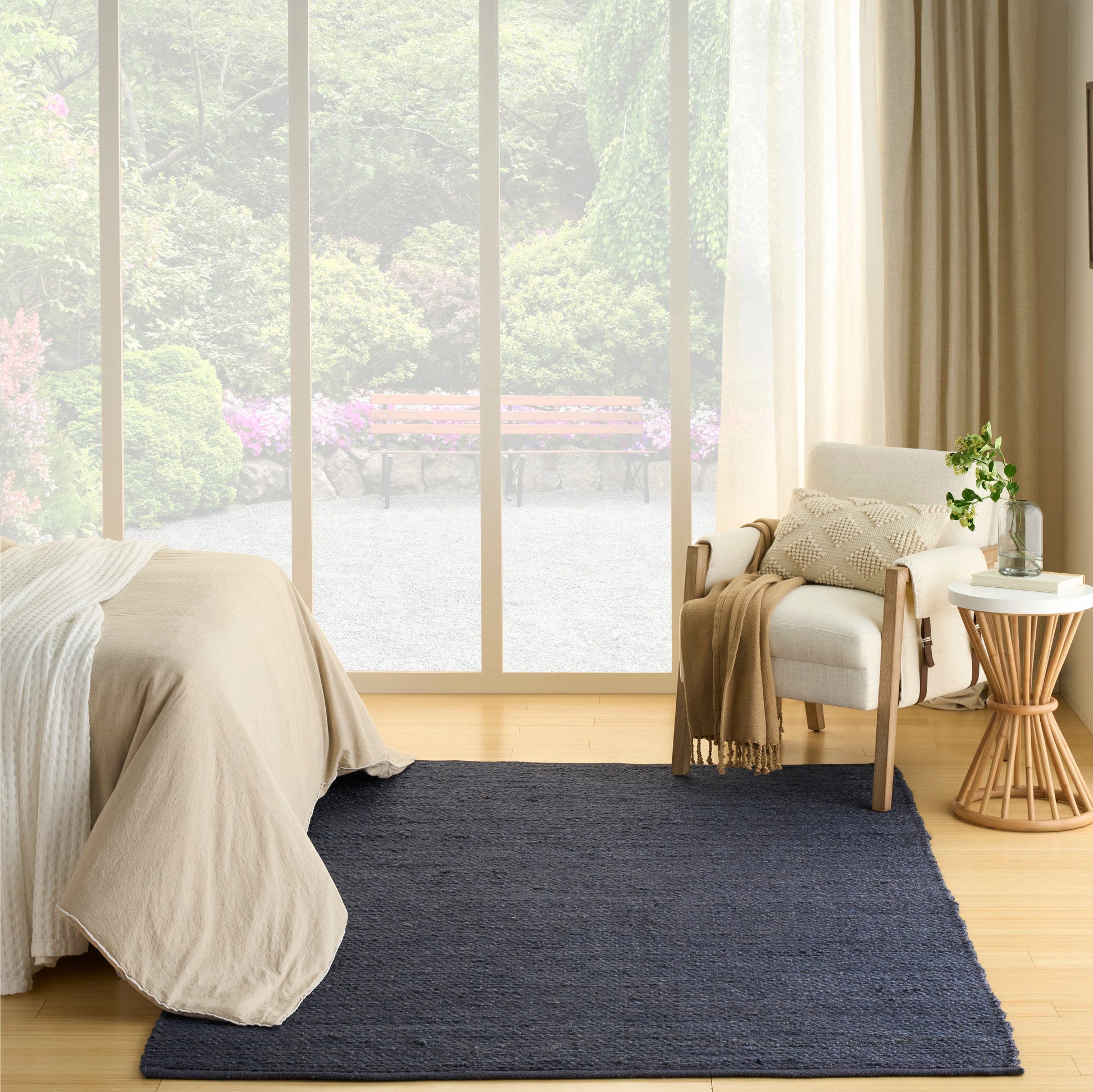 Cozy minimalist bedroom with navy rug, beige bedding, light wood floor, large windows, white armchair, side table and plant.