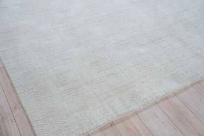 Light beige woven area rug with subtle texture, laid on pale wooden floor with visible edge.