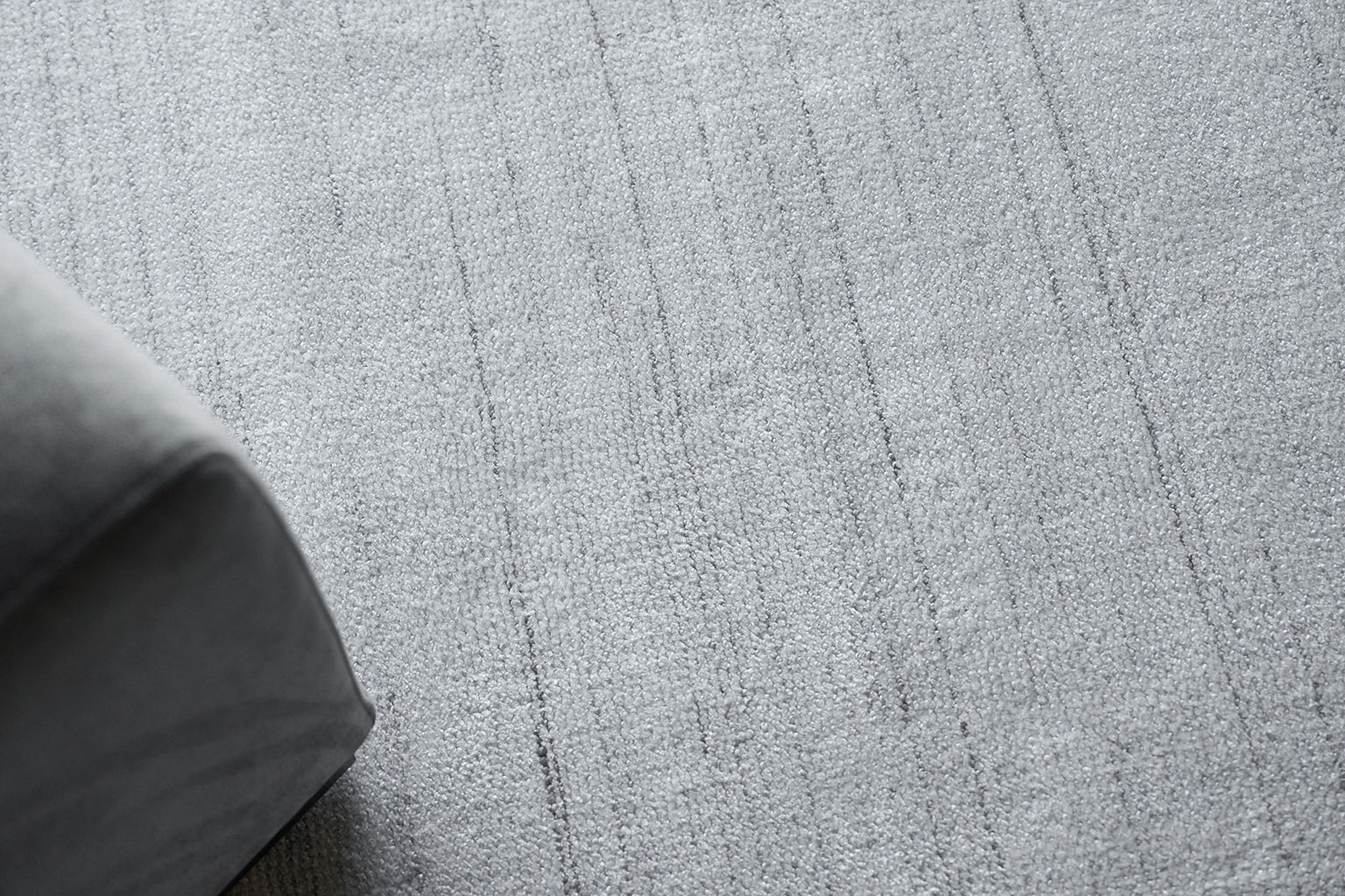 Close-up of light gray textured rug with subtle linear pattern; corner of gray upholstered furniture visible.