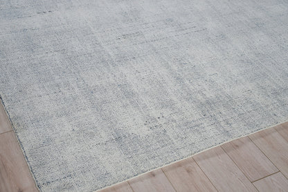 Light blue-gray hand loomed rug with a subtle distressed texture on pale wood flooring.