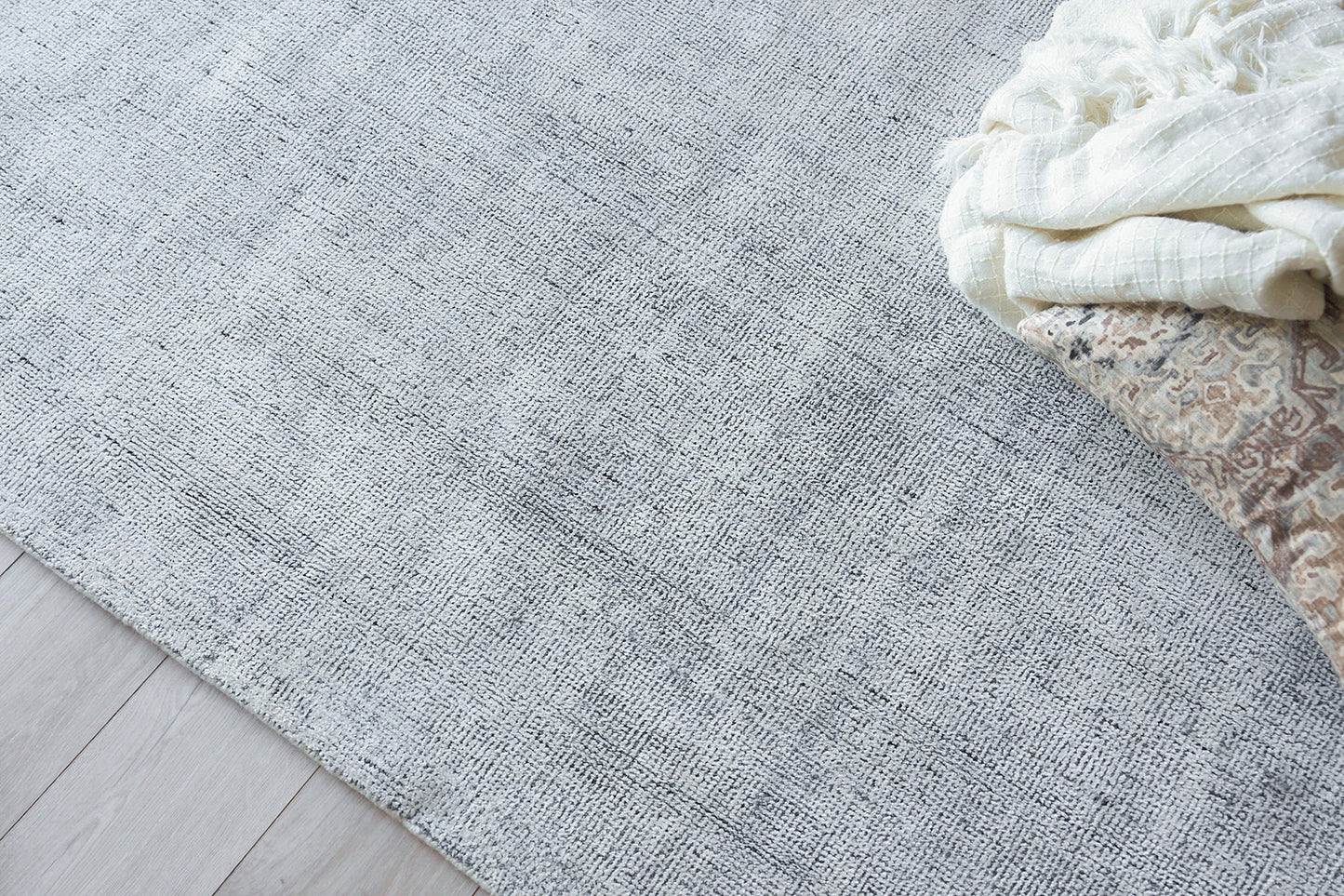 Light gray textured rug on pale wood floor with a folded white throw and patterned blanket in the corner.
