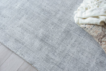 Light gray-blue textured area rug on pale wood floor with a white knit blanket and beige patterned throw.