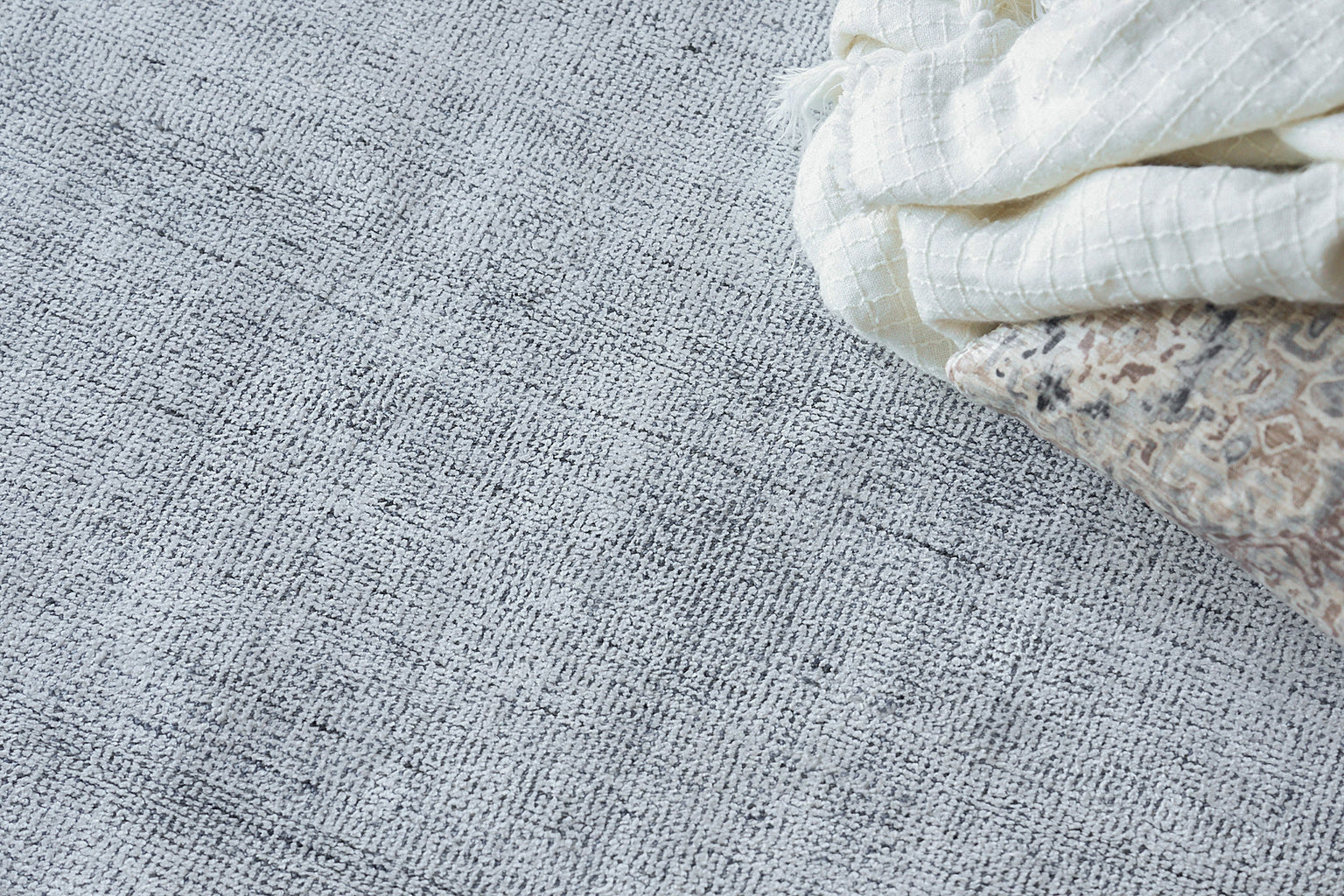 Gray textured rug with a white folded towel draped over a beige patterned cushion.