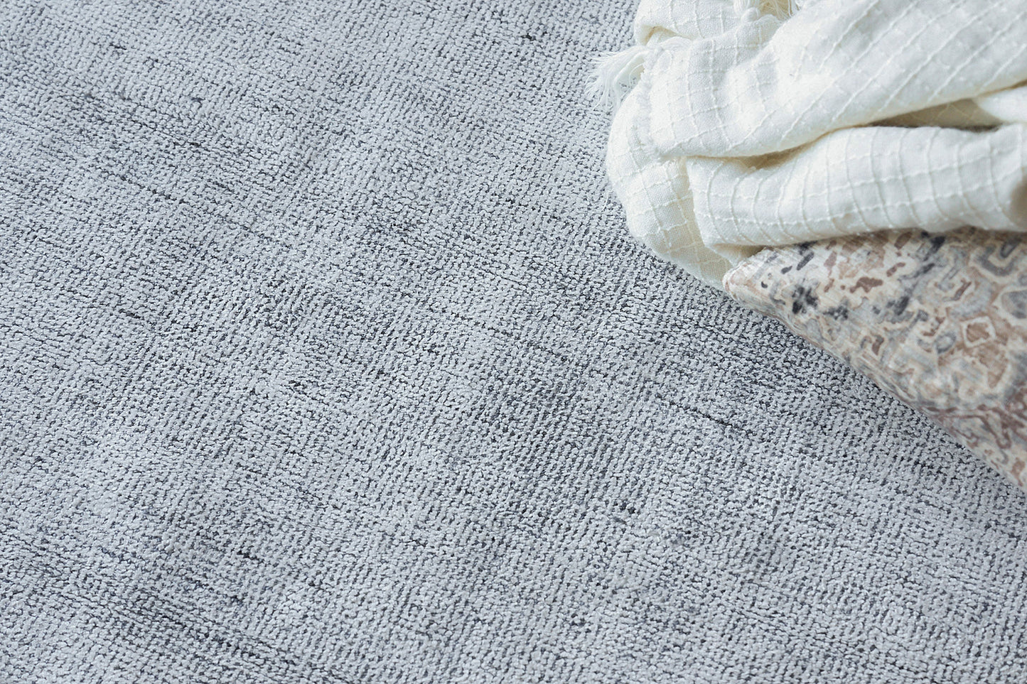 Light gray woven rug with cream folded towel & beige patterned fabric in the corner.