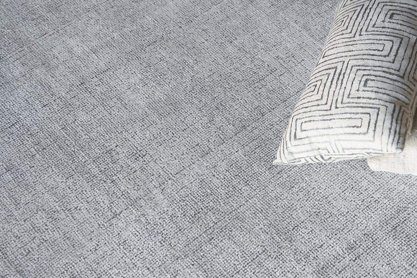 Gray textured rug with a cream rectangular pillow featuring a black geometric maze pattern in the corner.