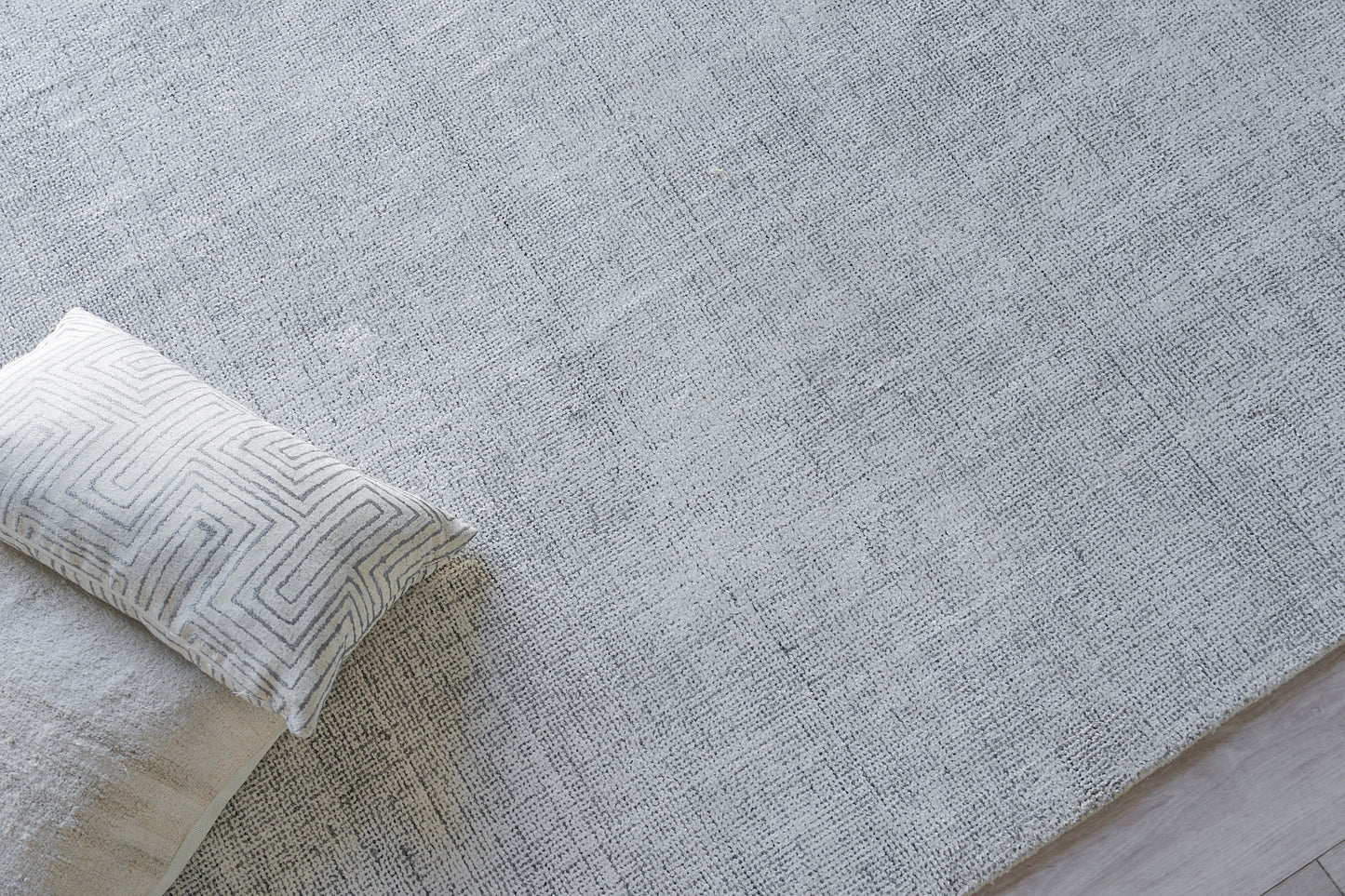 Light gray textured rug with a white geometric-patterned bolster pillow and cream throw at edge.